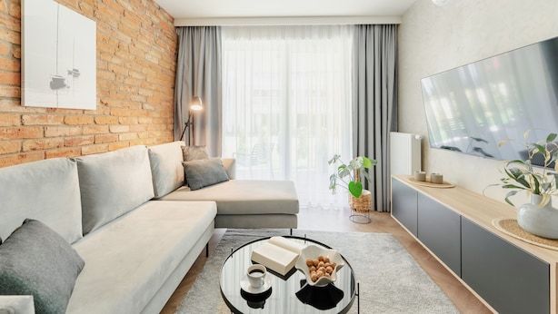 A modern living room with a sofa, a coffee table with books and a bowl of nuts, a TV, and a brick wall. - Apartments & SPA Posesja Plażowa Grzybowo by POSESJA APARTAMENTY 4a I (Grzybowo)
