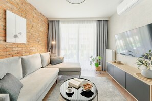A modern living room with a sofa, a coffee table with books and a bowl of nuts, a TV, and a brick wall. - Apartments & SPA Posesja Plażowa Grzybowo by POSESJA APARTAMENTY 4a I (Grzybowo)
