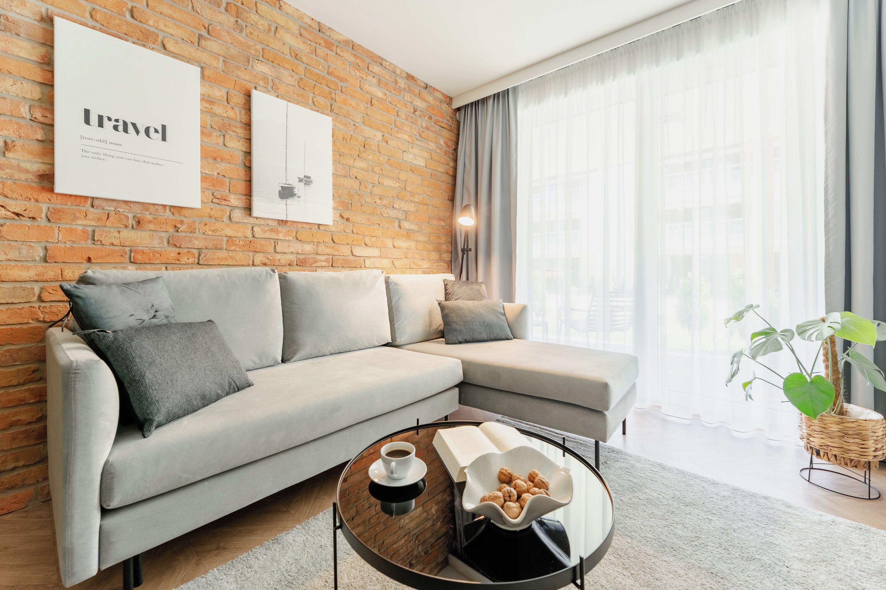 Un salon moderne avec un mur en briques, un canapé gris, une table basse avec un livre et des pâtisseries, et une grande fenêtre avec des rideaux.