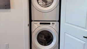 A stacked washer and dryer unit in a laundry room with a white door and a small wall-mounted artwork. - Charming 1BR Retreat - Walk to Gruene Hall + Upscale Amenities (New Braunfels)