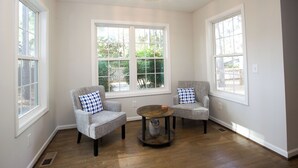 Una habitación con dos sillones grises, una mesa de centro redonda y amplios ventanales con marcos blancos.