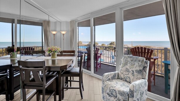 A balcony with a view of the beach, a dining table with chairs, and a floral armchair. - Serene Oceanfront 3 bedroom condo with breathtaking views (North Topsail Beach)