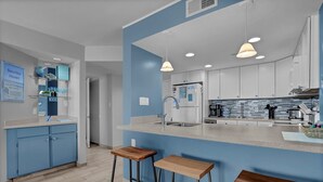 A modern kitchen with a blue accent wall, white cabinets, and a central island with bar stools. - Serene Oceanfront 3 bedroom condo with breathtaking views (North Topsail Beach)