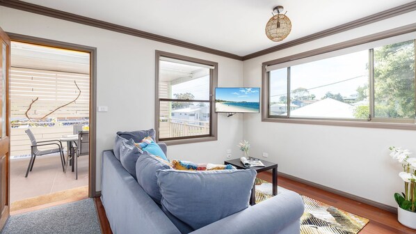 Una sala de estar moderna con un sofá gris, un televisor montado en la pared y grandes ventanales que ofrecen vista a la playa.