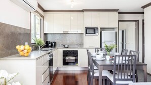 A modern kitchen with white cabinets, a built-in oven, a microwave, and a dining area with a table and chairs. - Lionel Gem – walk to Shoal Bay beaches and shops (Shoal Bay)