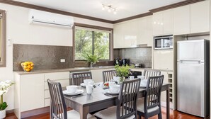 A modern kitchen with a dining table set for four, a refrigerator, and a window with a view of trees. - Lionel Gem – walk to Shoal Bay beaches and shops (Shoal Bay)
