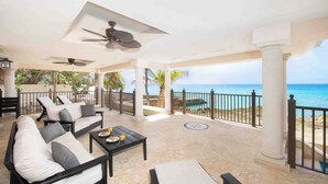 A covered outdoor seating area with white and gray cushioned chairs, a table with a plate of food and drinks, and a view of the ocean.