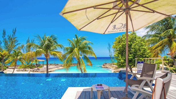 A wooden deck with a pool, lounge chairs, and an umbrella overlooking a beach with palm trees and clear blue water.