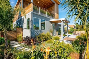 Ein modernes, zweistöckiges Haus mit Balkon, umgeben von einem gepflegten Garten mit verschiedenen Pflanzen und einer Holzterrasse.