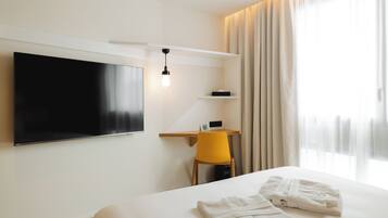 A modern hotel room with a large bed, a flat-screen TV mounted on the wall, a desk with a chair, and a window with curtains.