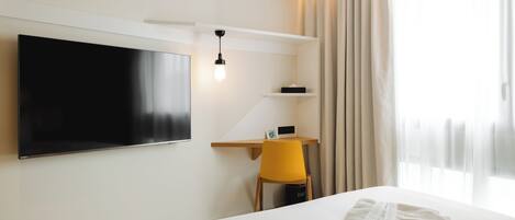 A modern hotel room with a large bed, a flat-screen TV mounted on the wall, a desk with a chair, and a window with curtains.