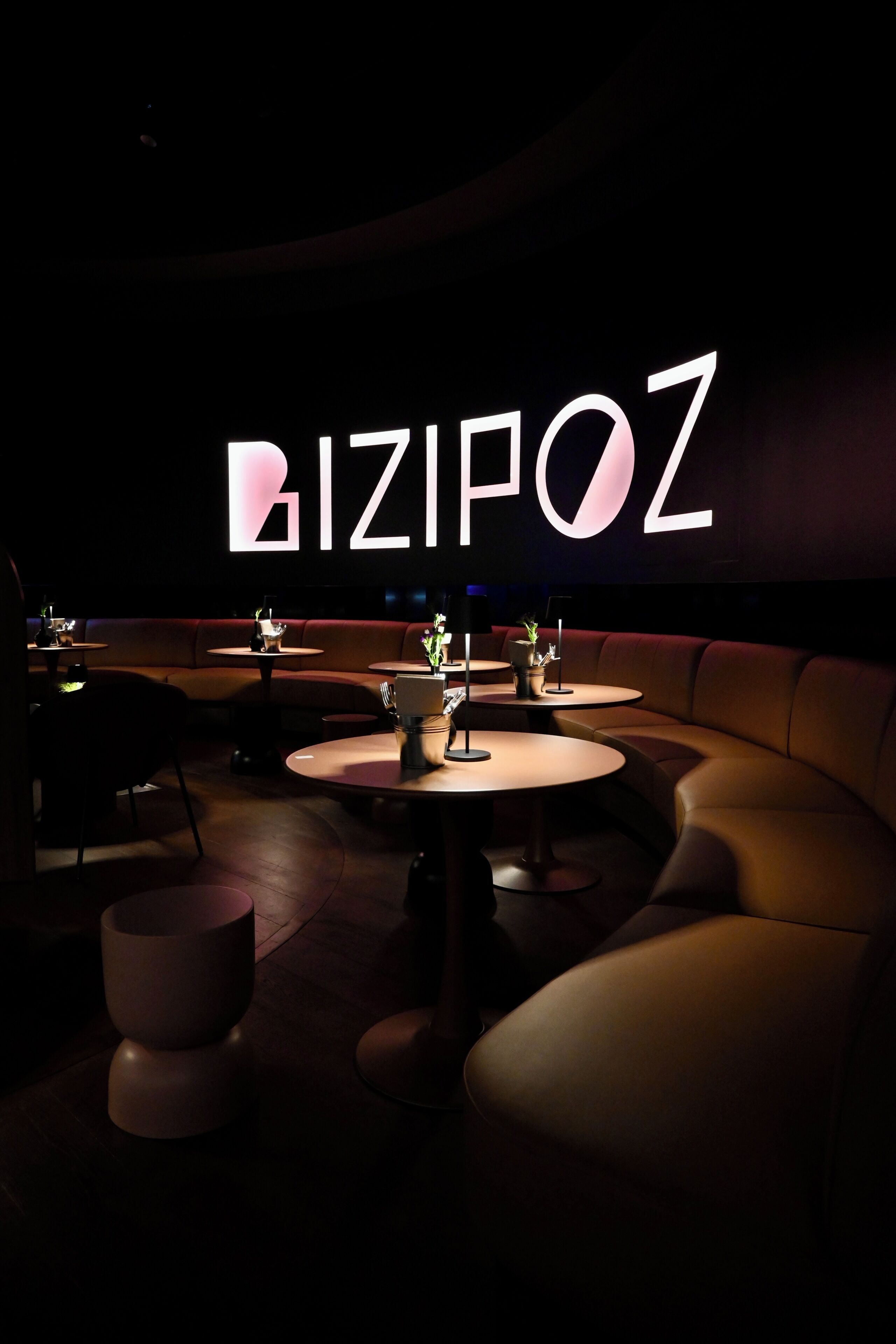 A dimly lit lounge area with a large illuminated sign reading 'BIZIPOZ,' featuring round tables and leather seating.