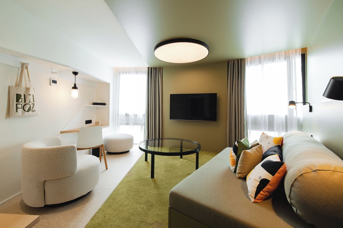 Una habitación de hotel moderna con sofá, sillón, mesa de centro de vidrio y televisor de pantalla plana montado en la pared.