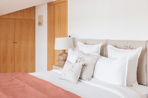 A neatly made bed with white and beige bedding, a wooden wardrobe, and a bedside table with a lamp.