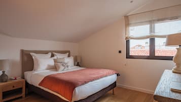 Een slaapkamer met een bed, nachtkastje, lamp en raam met uitzicht op gebouwen.