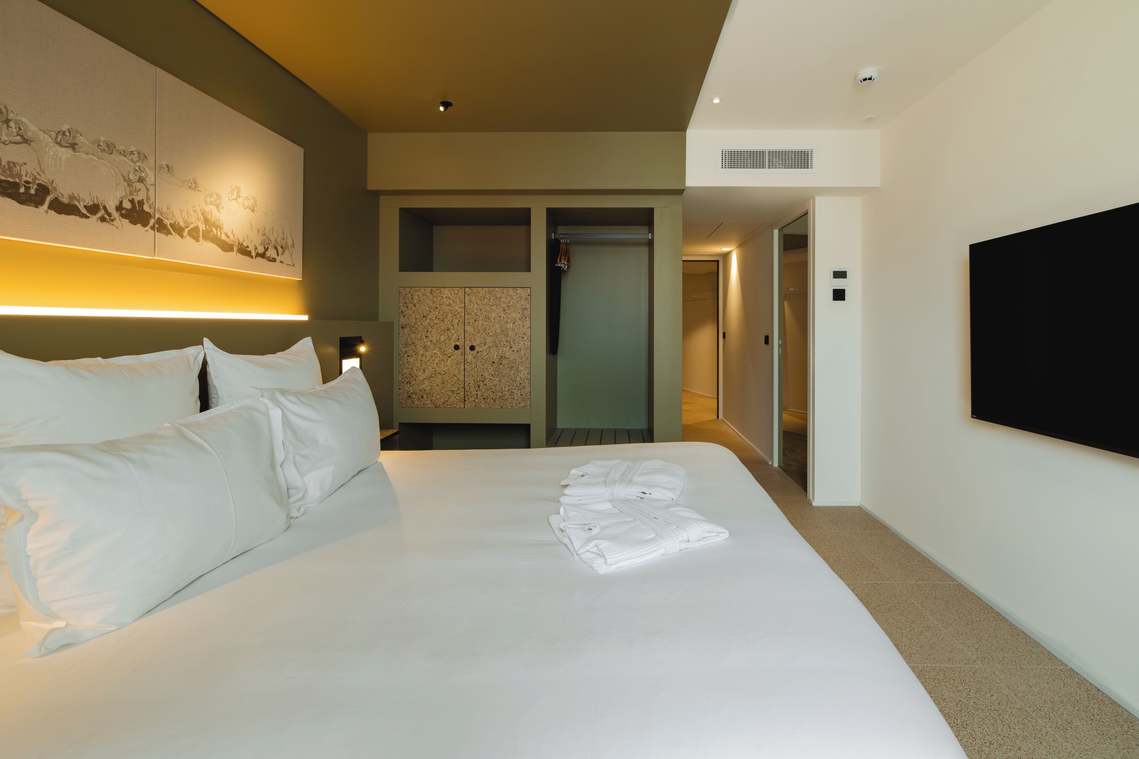 A hotel room with a large bed, a flat-screen TV mounted on the wall, a wardrobe, and a hallway leading to another room.