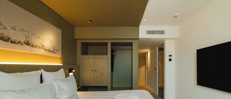 A hotel room with a large bed, a flat-screen TV mounted on the wall, a wardrobe, and a hallway leading to another room.