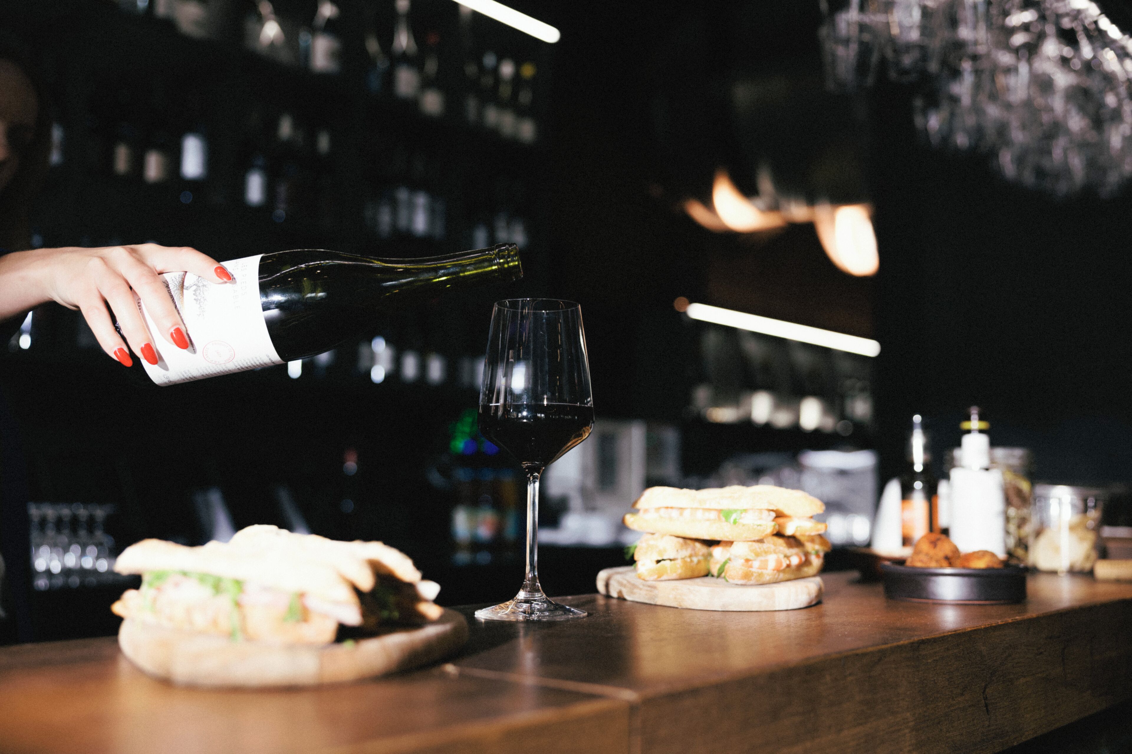 A bar setting with a wine bottle being poured into a glass, sandwiches on the counter, and various bottles in the background.