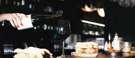 A bar setting with a wine bottle being poured into a glass, sandwiches on the counter, and various bottles in the background.