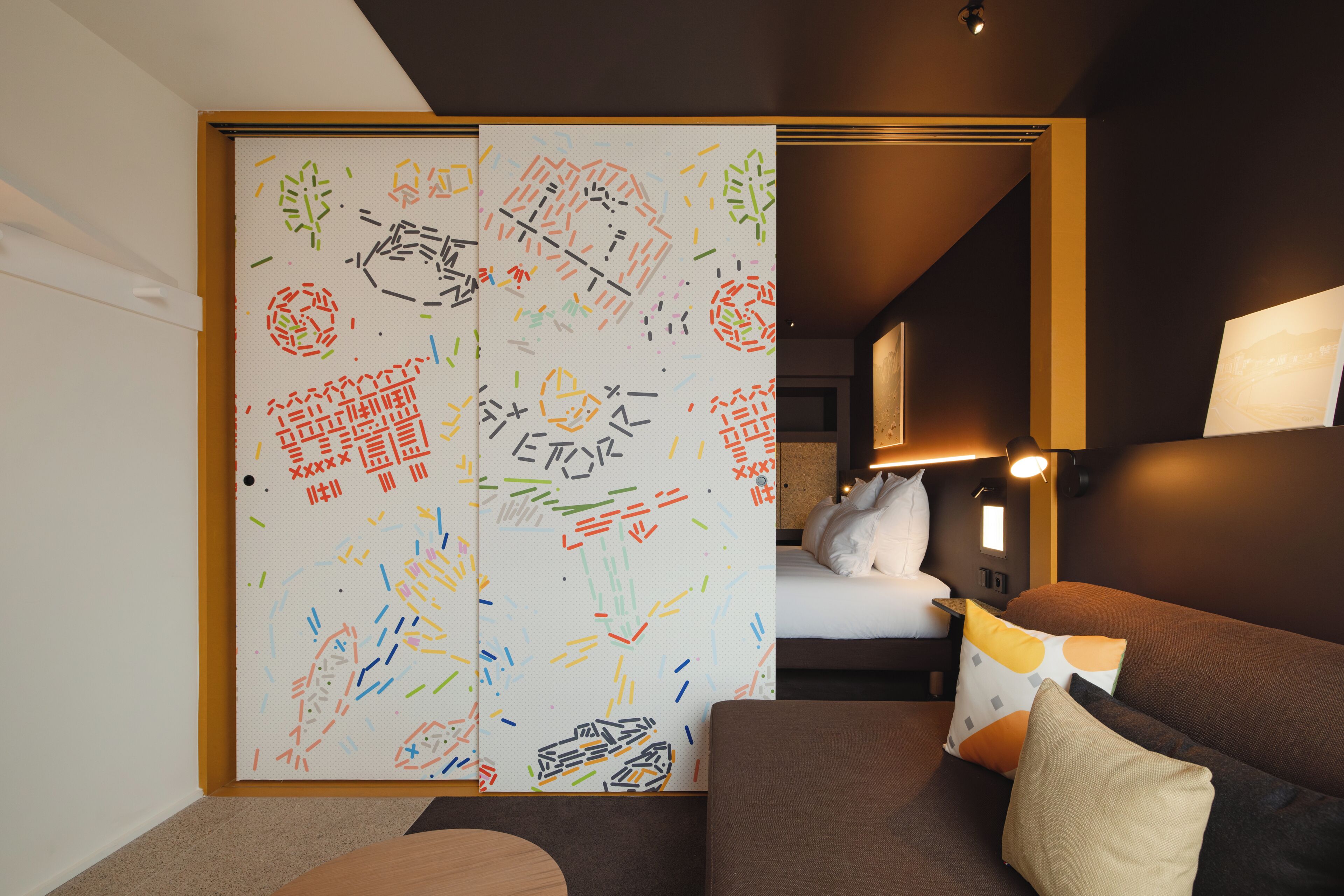 A hotel room with a bed, a brown sofa, a lamp, and a sliding door with colorful patterns.
