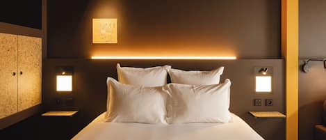 A hotel room with a large bed, bedside lamps, a headboard with integrated lighting, a small table, and a wardrobe.
