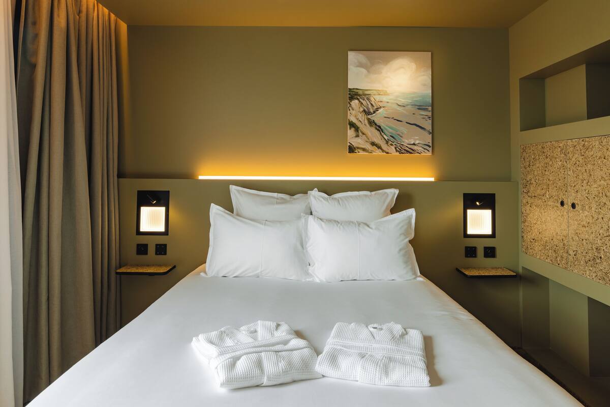 Habitación de hotel con una cama grande, dos mesitas de noche, una obra de arte en la pared y vistas a las montañas.