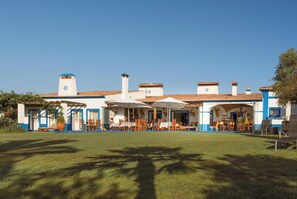Traditional Mediterranean-style building with rooms and a patio area, surrounded by a well-maintained lawn and shaded by trees.