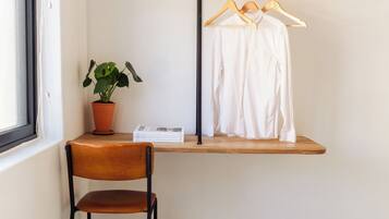 A room with a wooden shelf holding a white shirt and a hat, a potted plant, and a chair.