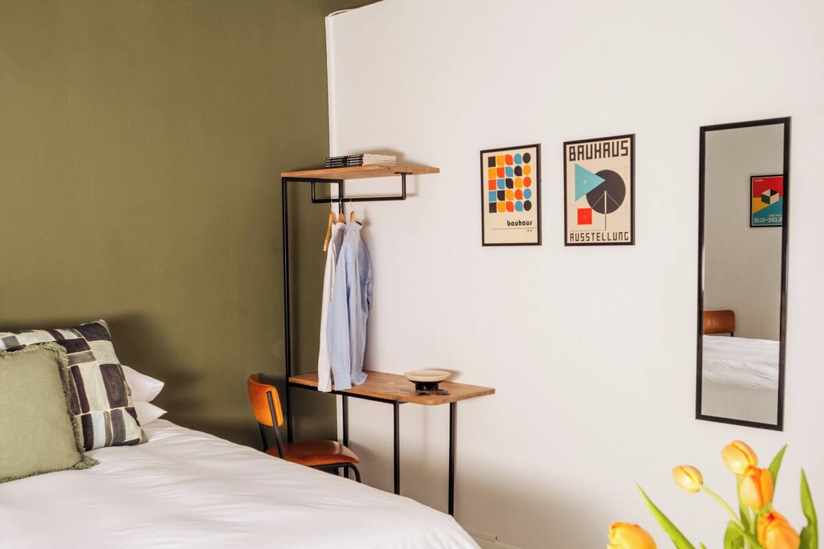A hotel room with a bed, a desk, a chair, a small table with a vase of tulips, and two framed pictures on the wall.