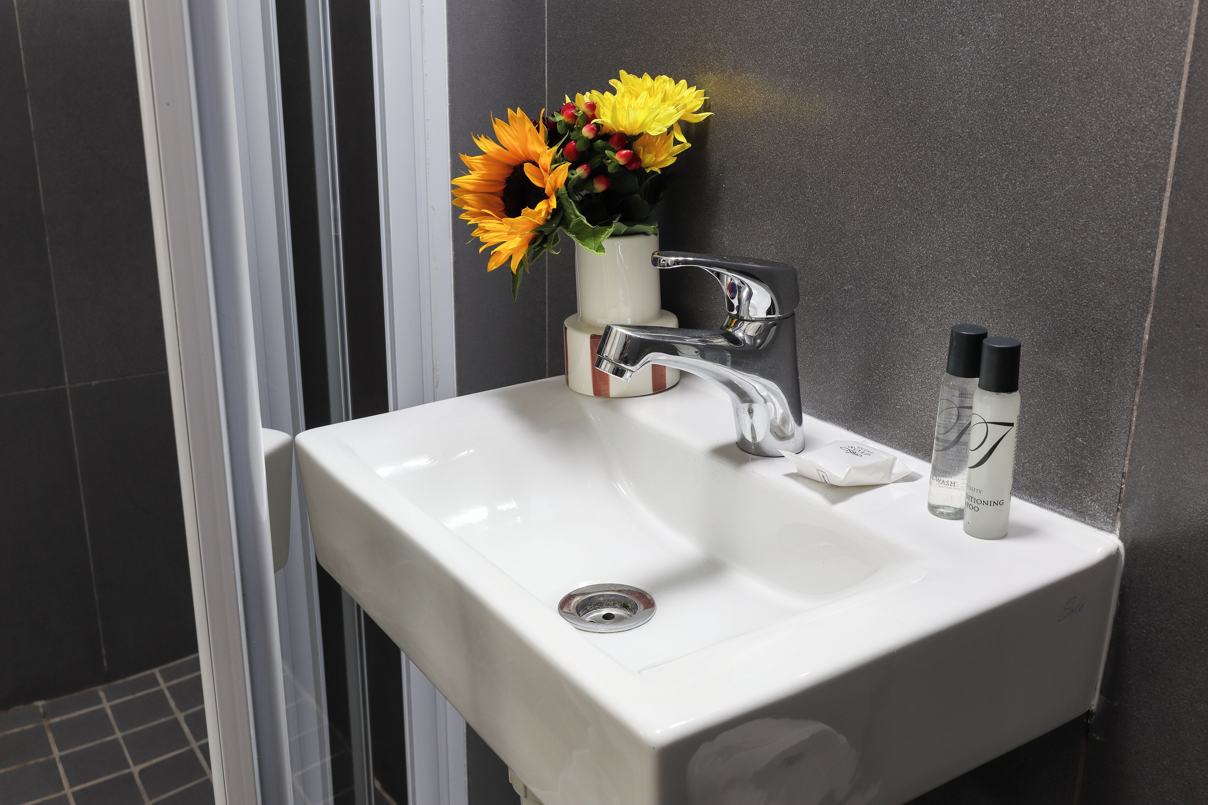 Un lavamanos de baño con un grifo moderno, un florero con flores amarillas y dos botellas de champú.