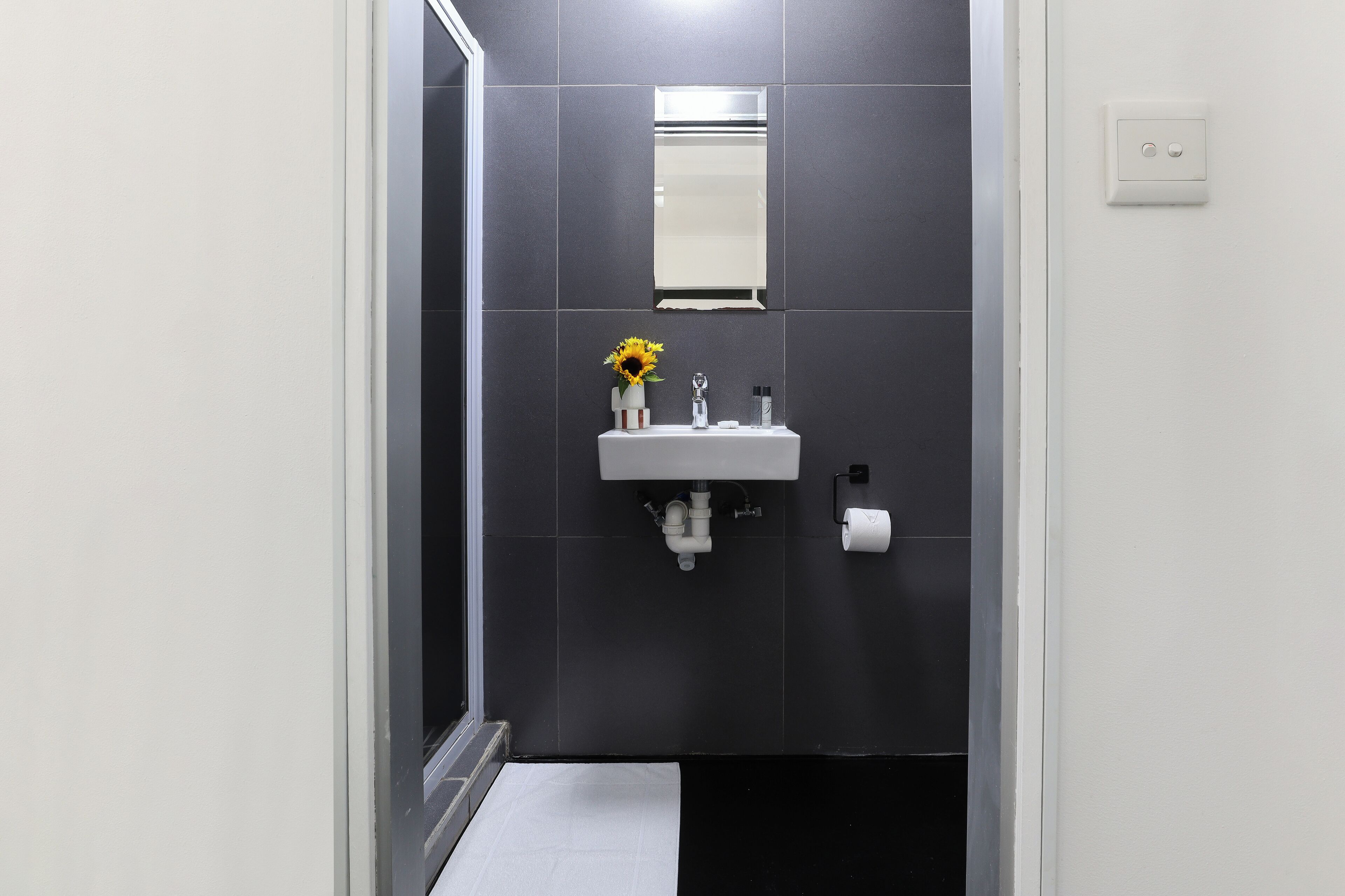 A modern bathroom with a white sink, dark tiles, a mirror, and a toilet paper holder.