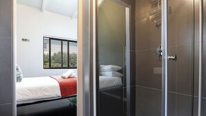 A modern hotel room with a bed, a shower area, and a window with blinds. - Fools Inn (Cape Town)