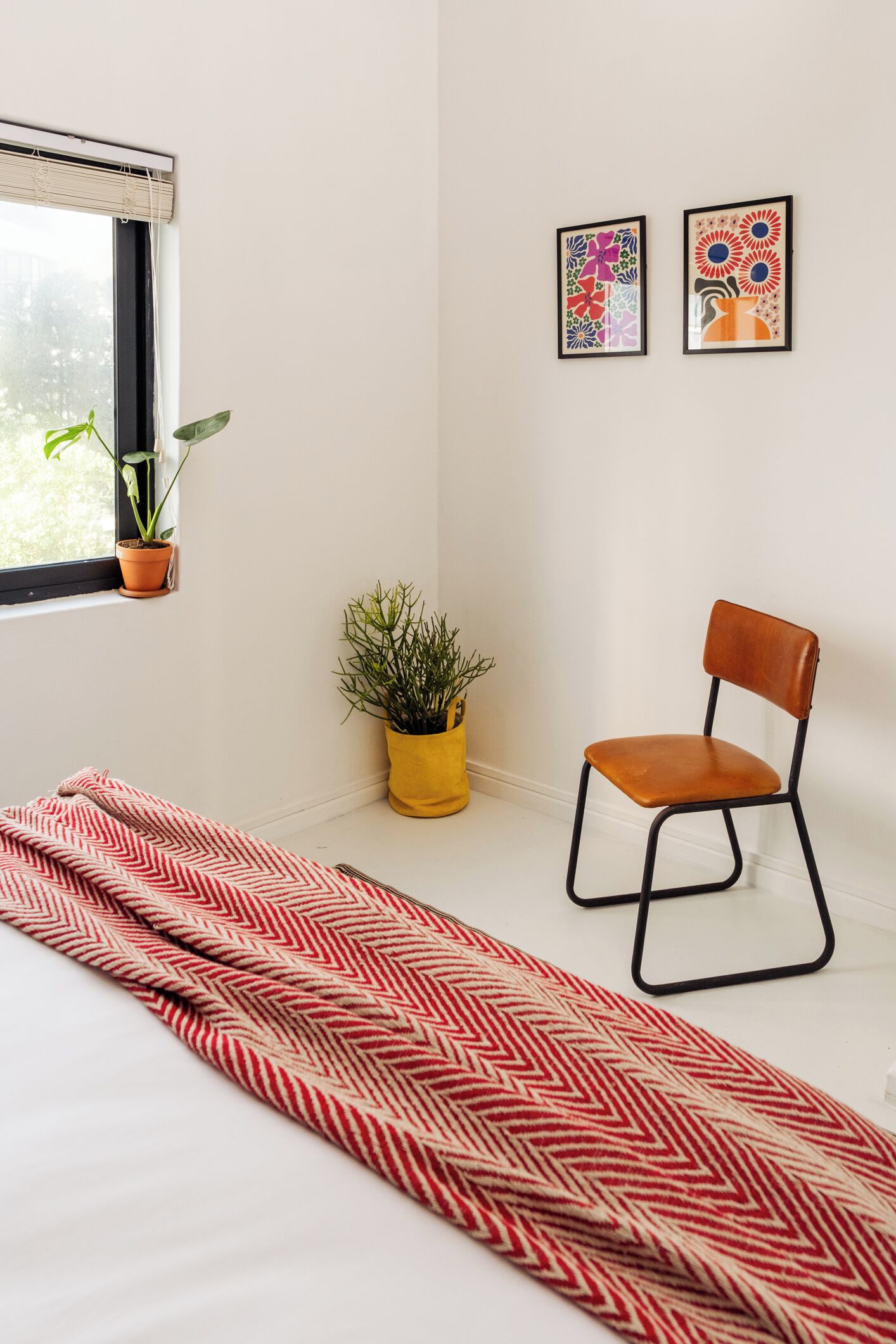 Quarto com cama, uma cadeira, duas plantas em vasos e duas obras de arte emolduradas na parede.