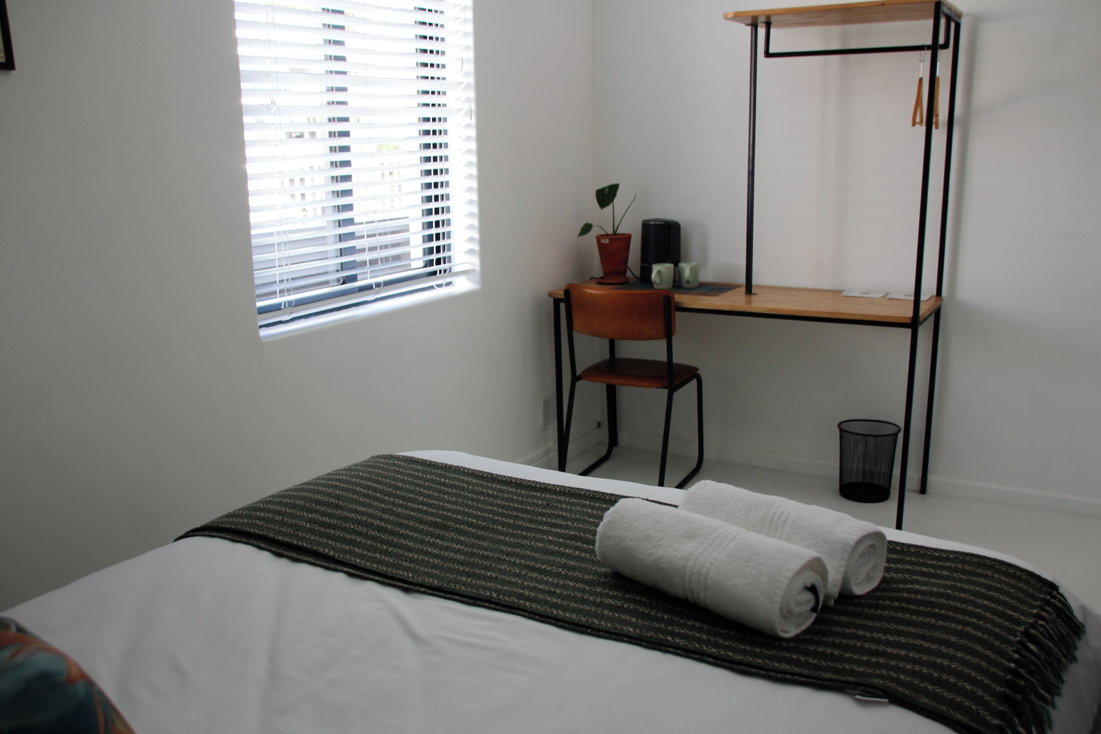 A bedroom with a bed, a desk, a chair, a window with blinds, and a small plant.
