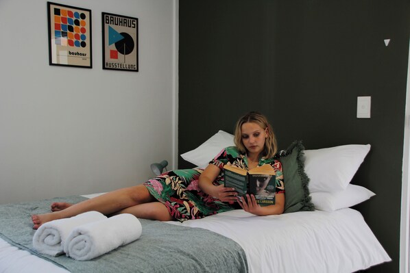 A person lying on a bed with a book, a pillow, and rolled towels. - Fools Inn (Cape Town)