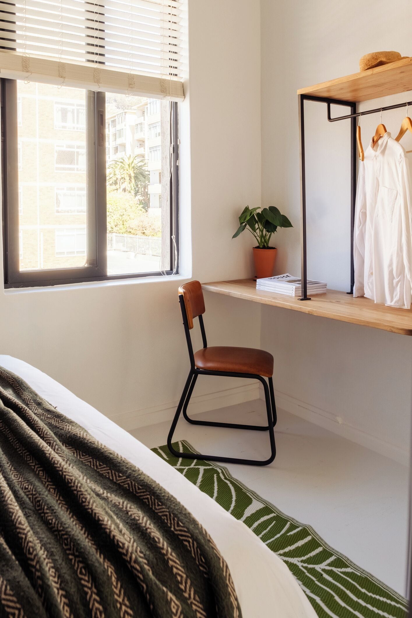 A bedroom with a bed, a chair, a shelf with clothes and a plant, and a window with blinds.