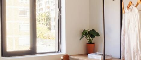 A bedroom with a bed, a chair, a shelf with clothes and a plant, and a window with blinds.