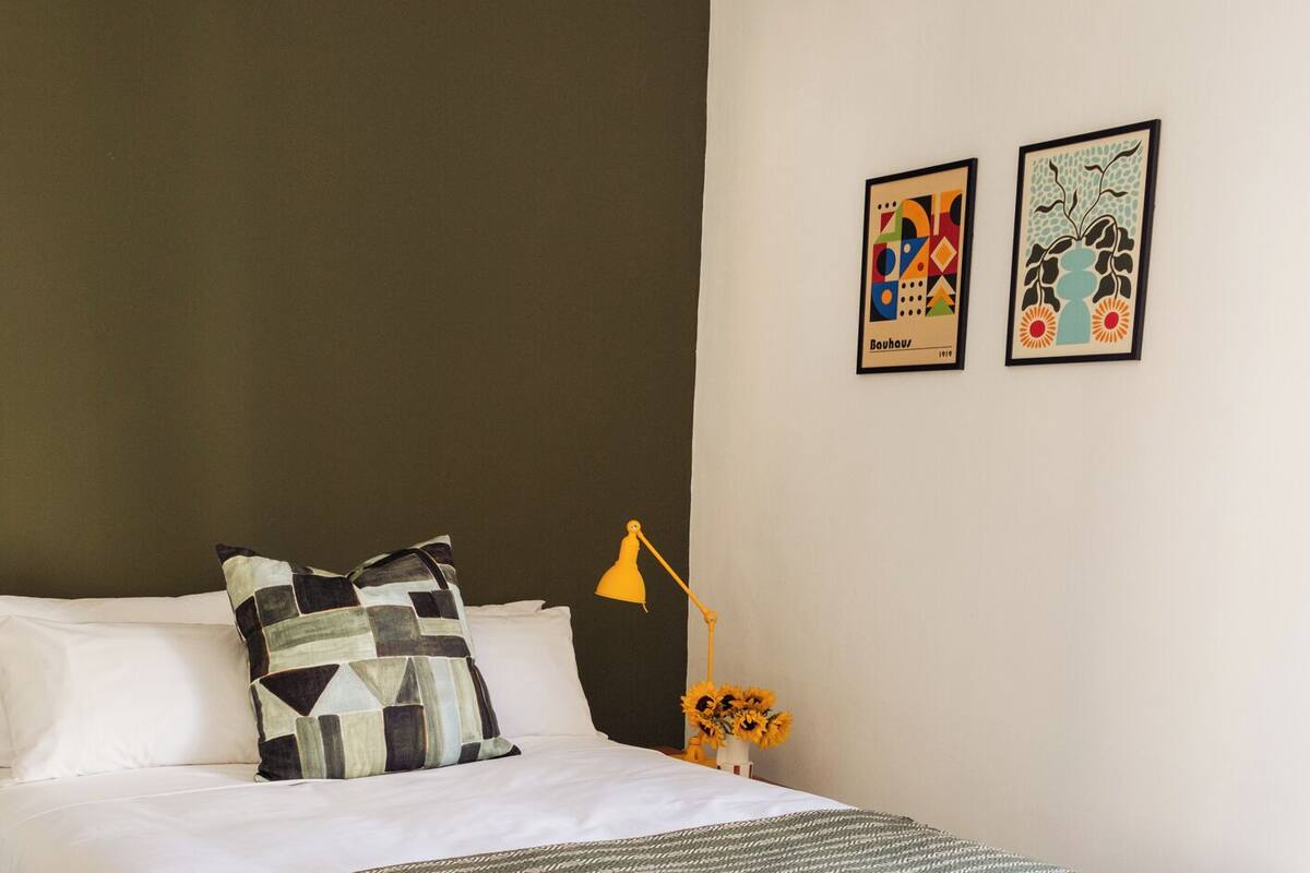 A neatly made bed with a patterned throw, a green leaf-patterned rug, and two wall art pieces.