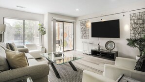 Un salon moderne avec une table basse en verre, un canapé, un téléviseur fixé au mur et une porte-fenêtre coulissante donnant sur un espace extérieur.