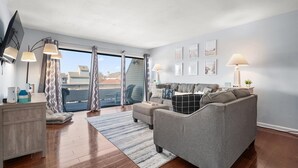 A modern living room with a grey sofa, a coffee table, and a TV on a stand. There are curtains, a rug, and framed pictures on the wall. - 2 bedroom, 2 bath, 28th bayside with pool and tennis court! (Ocean City)