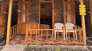 Un chalet en bois avec un toit vert, une véranda dotée de deux chaises en plastique blanches et une vue sur une montagne boisée.