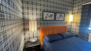A bedroom with a checkered wall pattern, a wooden headboard, a bed with blue bedding, a bedside table with a lamp, and two framed pictures on the wall. - Superior Double Room (Avellino)