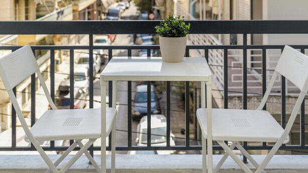Een balkon met twee witte stoelen en een klein tafeltje, met uitzicht op een smalle straat met gebouwen.
