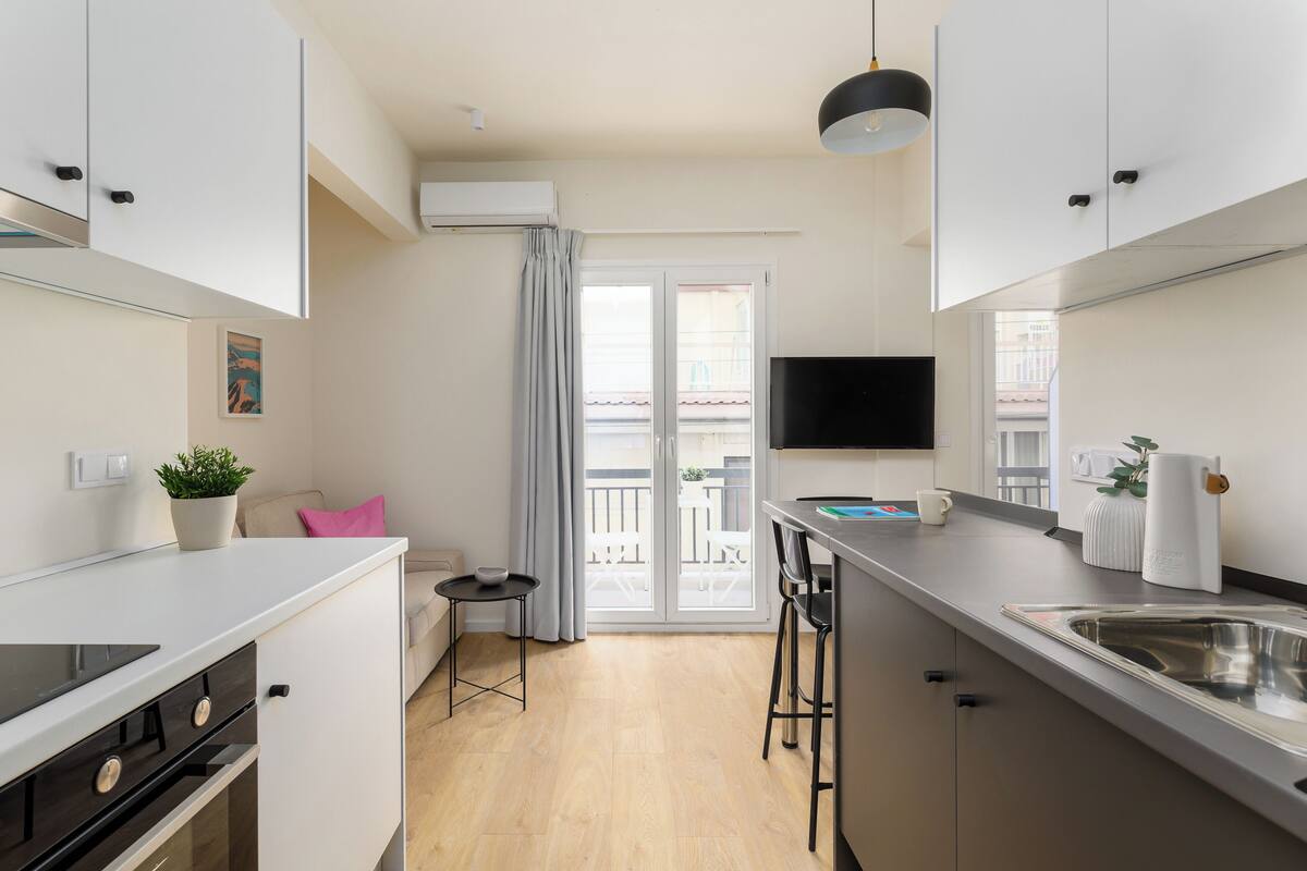 Una cucina moderna con piano cottura integrato, un'area pranzo con sgabelli da bar, una TV a schermo piatto e una porta scorrevole in vetro che dà su un balcone.
