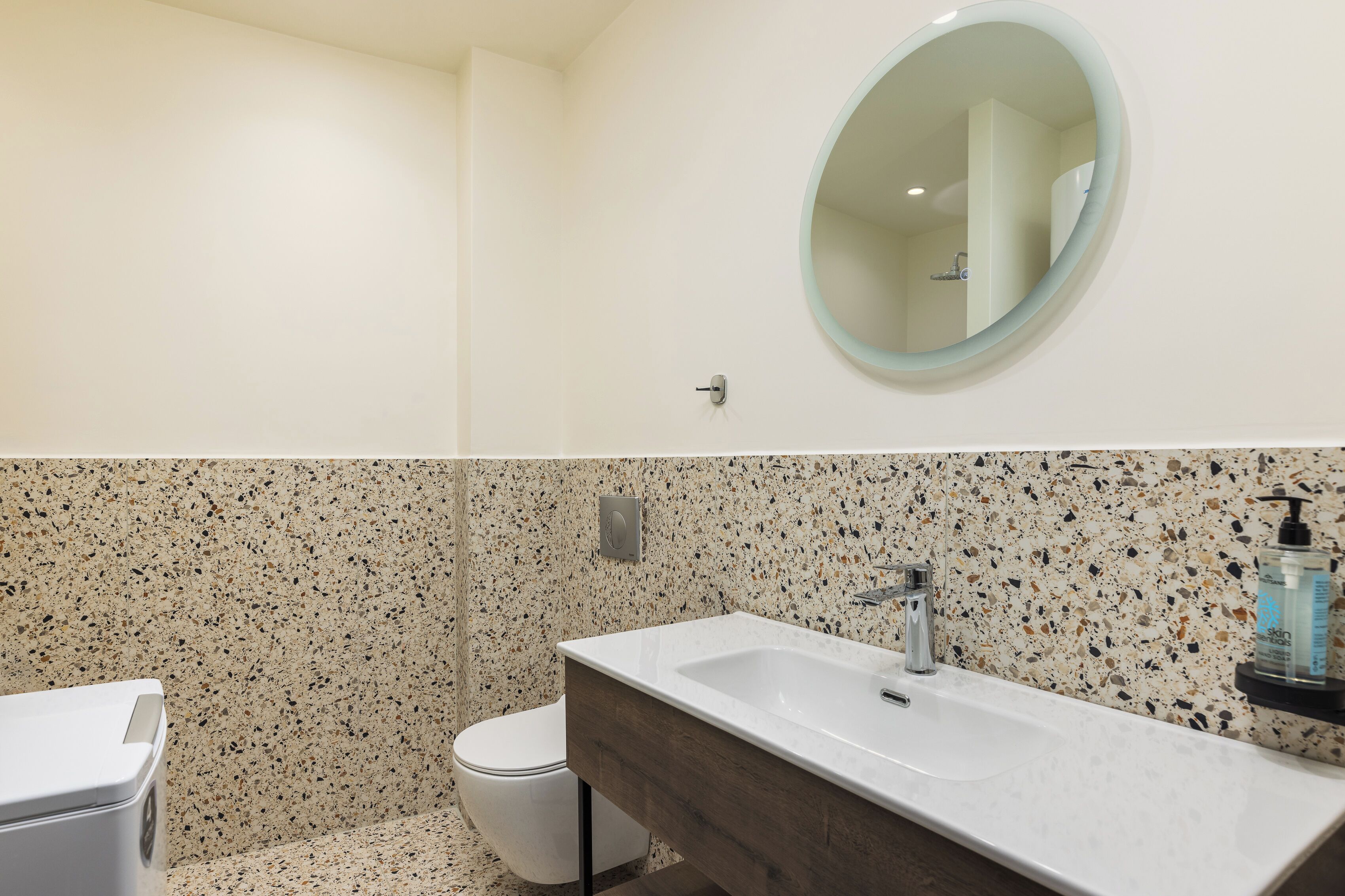 A bathroom with a white toilet, a white sink with a wooden vanity, a round mirror, and a speckled backsplash.