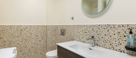 A bathroom with a white toilet, a white sink with a wooden vanity, a round mirror, and a speckled backsplash.