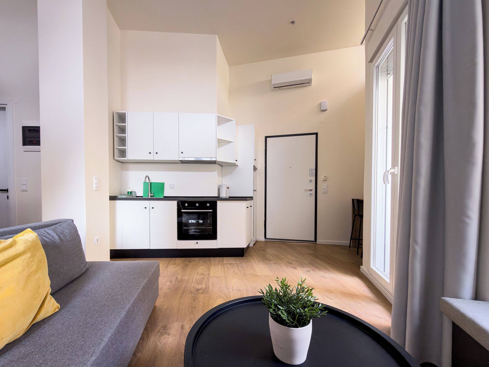 Ein kompakter Wohnraum mit Küchenzeile, einem Sofa mit gelbem Kissen, einer Topfpflanze und einem großen Fenster mit Vorhängen.