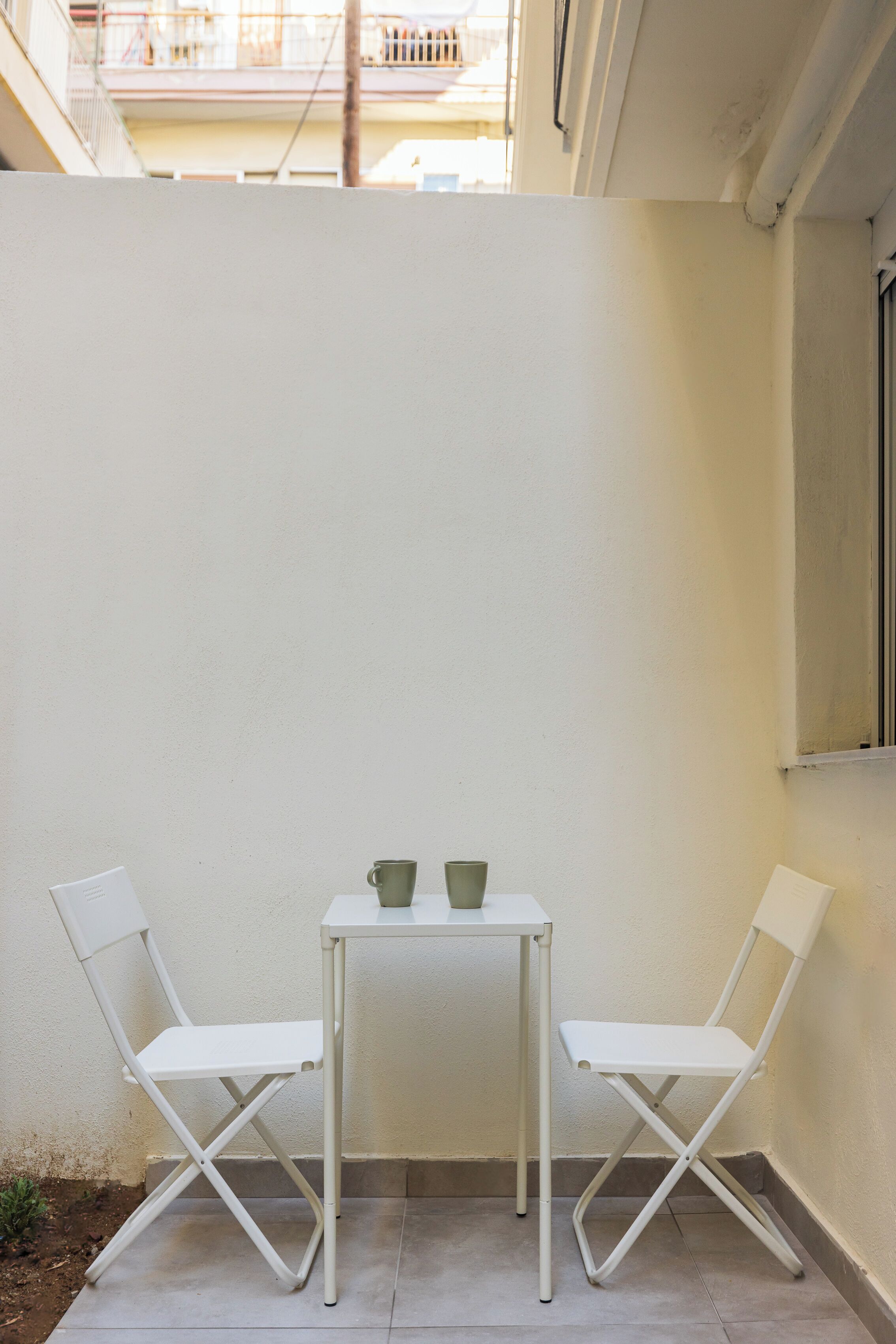 A small outdoor seating area with two white chairs and a table, a window, and a balcony in the background.