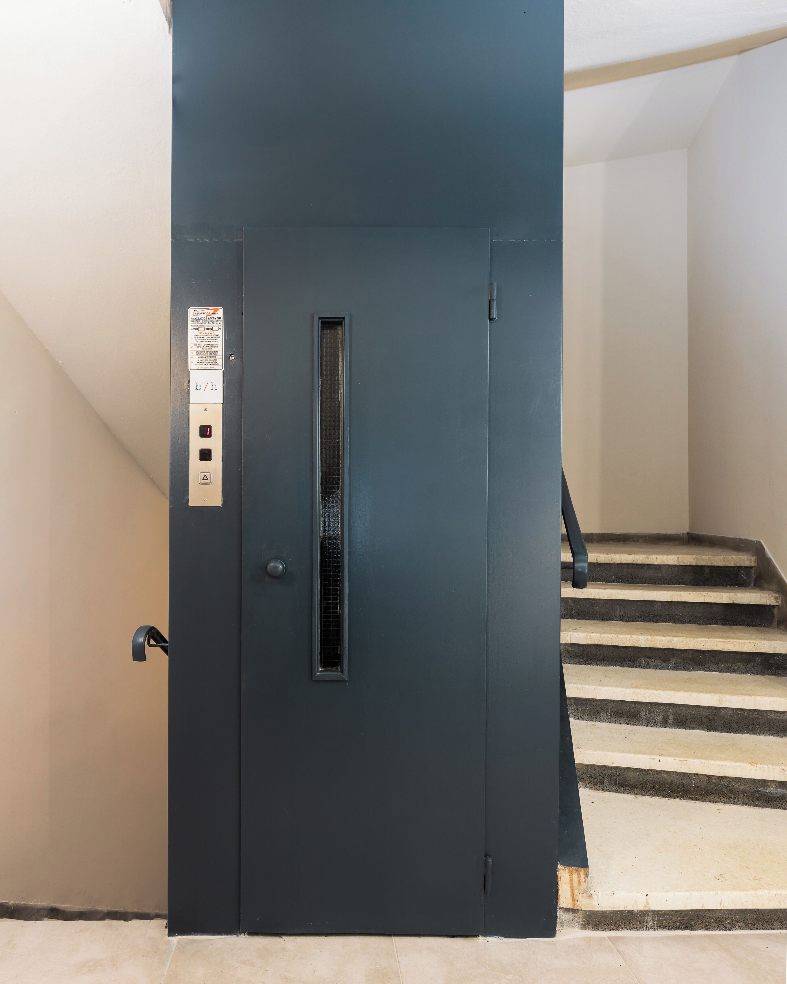 Une porte d’ascenseur bleu foncé, dotée d’un panneau de commande, d’une grille de ventilation verticale et d’une main courante sur un escalier.