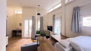 Een moderne hotelkamer met een houten vloer, een bed met witte lakens, een bureau met een plant, een stoel, een aan de muur bevestigde televisie en grote ramen met gordijnen.